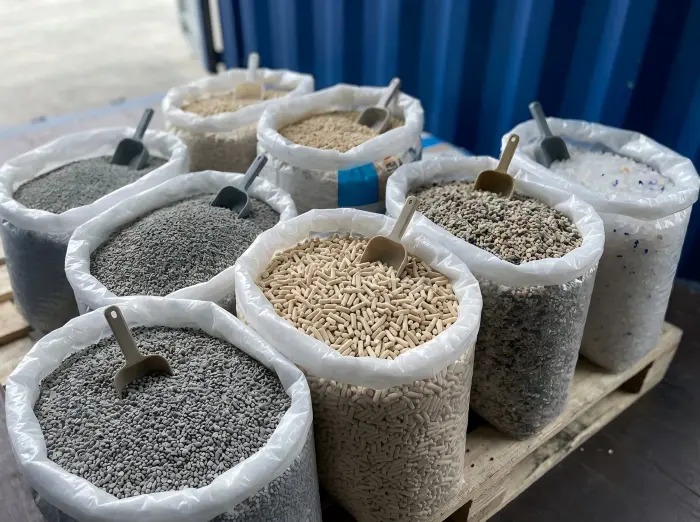 Different types of cat litter including bentonite ore, plant tofu pellets, and silica gel crystals in open bags on a wooden pallet.